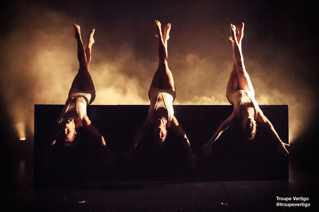 Three @troupevertigo dancers upside down with legs crossed and arms extended.